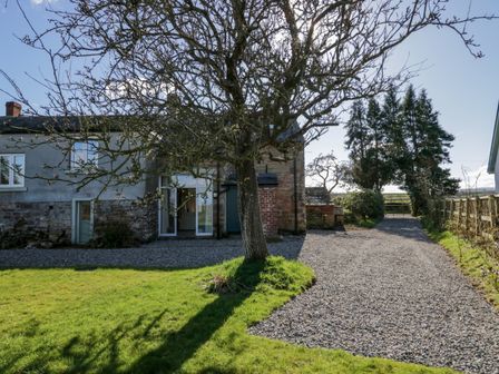 A gravel driveway with a tree on a lawn beside a stone and brick house at The Roost in Cumdivock near Dalston