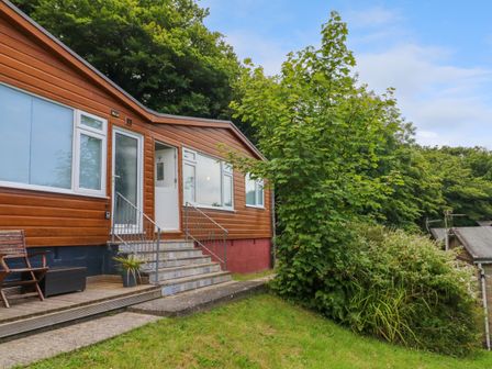 An exterior view of a wooden house with stairs leading to a white door and a small deck with chairs and potted plants surrounded by grass and trees at 4 The Park in Kilkhampton