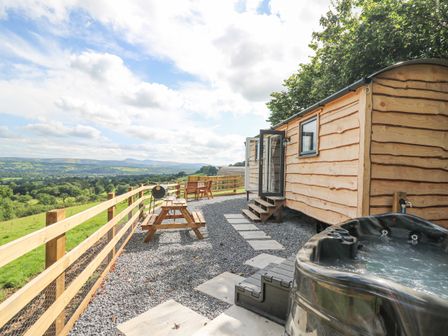 An outdoor area with a wooden cabin a hot tub wooden picnic table chairs and a barbecue with a scenic countryside view at Shepherd's Cwtch in Cilycwm near Llandovery
