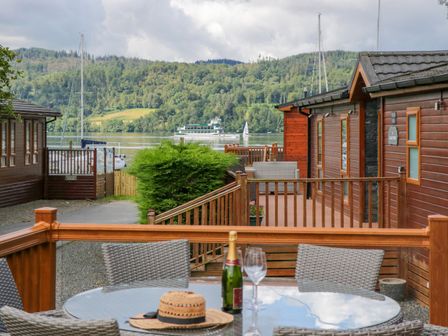 A waterside patio with a round glass table set with a bottle of champagne and hat between wooden lodges at Waterside Lodge 55A Bowness White Cross Bay near Windermere
