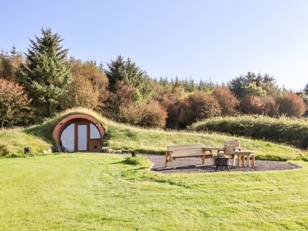 An earth house with a round door surrounded by grass and wooden outdoor furniture in a clearing at Rhosydd in Cyffylliog near Ruthin