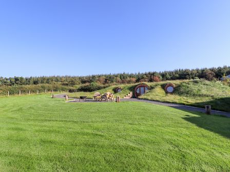 A grassy outdoor area with wooden chairs and table near round door homes built into a hillside at Weirglodd in Ruthin
