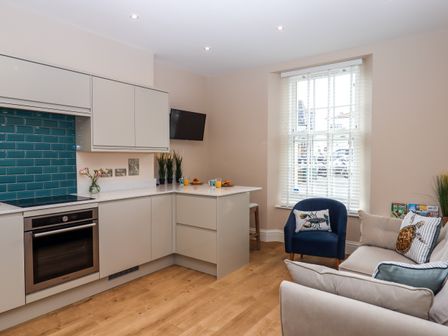 A kitchen area with beige cabinets a built in oven teal tile backsplash and a sitting area with sofa and blue chair at 1 Sea Otter House in Teignmouth