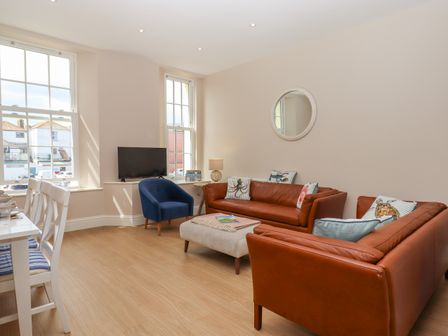 A living room with brown leather sofas a blue armchair a beige ottoman a TV on a stand and a dining table with white chairs at 2 Sea Otter House in Teignmouth