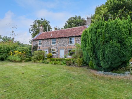 A stone cottage with a red tiled roof surrounded by a garden with bushes and a large tree at West View in Feltwell