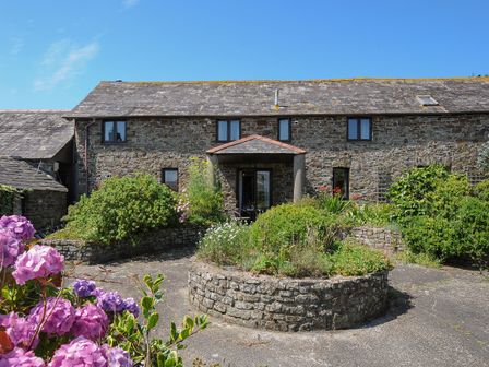 A stone building with a slate roof and a circular stone planter with greenery and flowers in front at West Lee in Bude