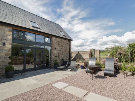 A stone building with large glass doors and windows beside a patio with lounge chairs and metal fire pit at The Hay Barn in Huntley