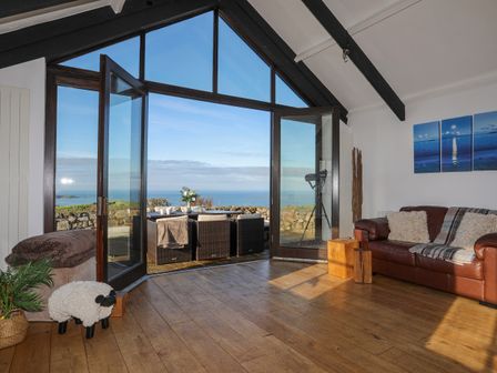 A living room with a leather sofa and open glass doors leading to an outdoor seating area with an ocean view at Tai Ffolt in Nefyn