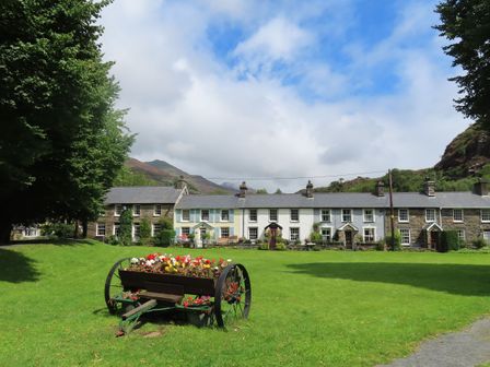 A row of terraced houses behind a green lawn with a flower-filled cart in the foreground at Cottage on the Green in Beddgelert