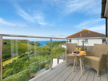A balcony with two wicker chairs and a small table with food and drinks overlooking green hills and the sea at Rowena in Mullion