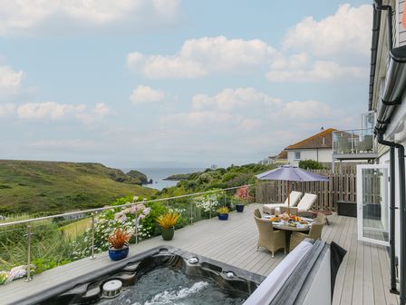An outdoor deck with a hot tub a table with four chairs and an umbrella two lounge chairs and plants overlooking a grassy hillside and sea at Morwenna in Mullion