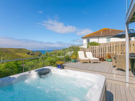 An outdoor deck with a hot tub two lounge chairs and a dining table with an umbrella overlooking a grassy hillside and ocean at Verity in Mullion