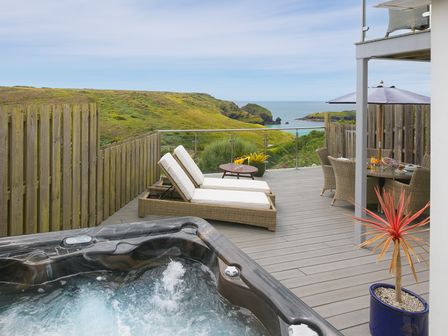 An outdoor deck with a hot tub two lounge chairs dining table with umbrella and ocean view at Daphne in Mullion