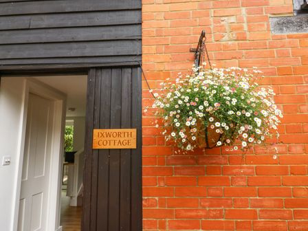 An open door with a wooden sign reading Ixworth Cottage and a hanging basket of flowers on a brick wall at Ixworth Cottage in Little Woodford near Ottery St Mary