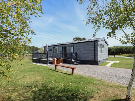 An exterior view of a single-story cabin with a deck and outdoor seating surrounded by grass and trees at Sunset Lodge in Woodhall Spa