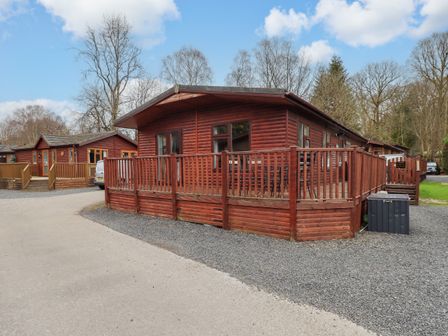 A wooden cabin with a fenced deck and outdoor furniture in a wooded area at Brightwater Lodge White Cross Bay near Windermere