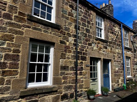 A stone cottage exterior with multiple white framed windows a green door and a blue door at Aln Cottage Alnmouth