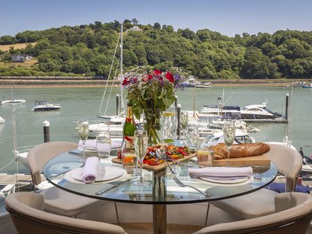 A glass dining table set with plates glasses bread and flowers overlooking a marina at 21 Dart Marina in Dartmouth