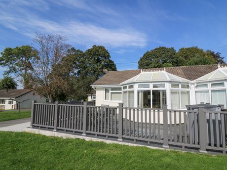A single-story house with a patio and gray fence surrounded by grass and trees at Trem-Yr-Afon in Aberaeron