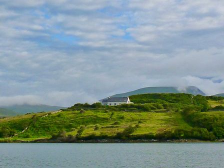 A house on a green hill next to a body of water with mountains and cloudy sky in the background at Lighthouse Hill in Tiernaur near Mulranny