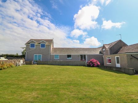 A house with a large grassy lawn and two white chairs near a flower bed at Gwynlys in Cemaes Bay