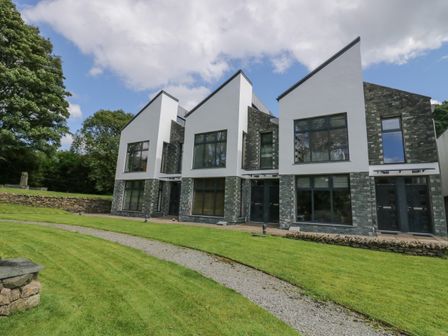 A row of modern houses with large windows and dark stone and white walls surrounded by grass and trees at 12 Blue Hill Park in Ambleside