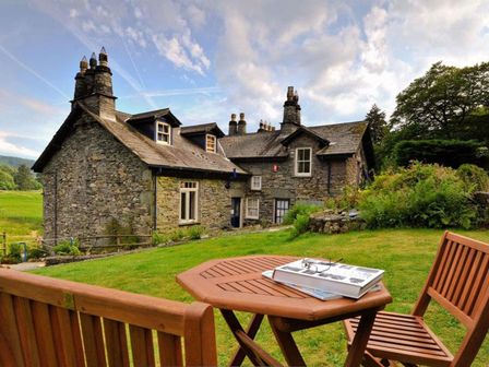 A stone house with multiple chimneys seen from a garden with wooden chairs and table at Riverbank At Stepping Stones in Rydal