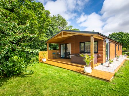 A wooden lodge with a covered porch and outdoor seating surrounded by grass and trees at Maple Lodge in Angarrack