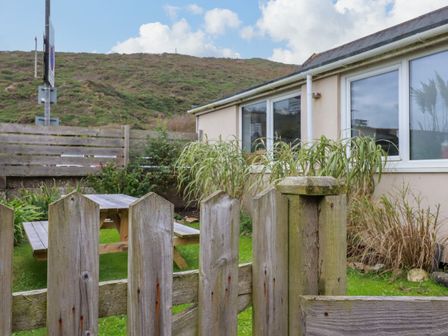 A wooden fence gate opening to a garden with a picnic table next to a house with windows at 1 Mill Cottages in Porthtowan