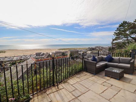 A patio with a wicker sectional sofa and glass table overlooking a beach town and the sea at Tonnau Yr Oleu or Waves of Light in Barmouth