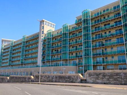 A large apartment building with green-tinted glass balconies and cylindrical glass elevator shafts along a street at Manhattan Beach in Castletown Portland