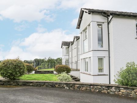 The exterior of a white house with bay windows a stone wall bushes and a paved driveway at Ramblers Lodge in Bowland Bridge near Bowness-On-Windermere Lake District