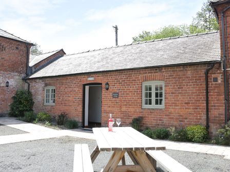 An outdoor area with a wooden picnic table and a bottle with a glass in front of a red brick building with two windows and an open door at Woodside Cottage in Kerry