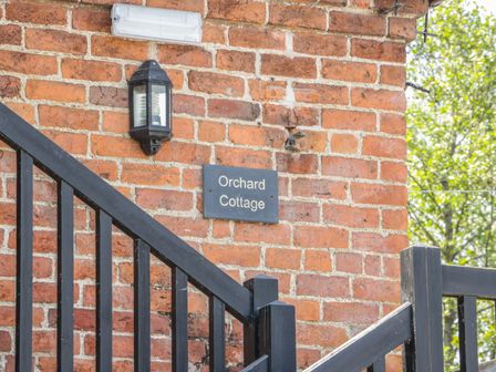 A brick wall with a black light fixture and a sign reading Orchard Cottage next to a black wooden fence with trees in the background at Orchard Cottage Kerry