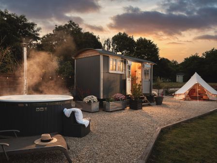 An outdoor area with a hot tub and lounge chair next to a small cabin and a lit tent at sunset at Stargazer in St Erme near Truro