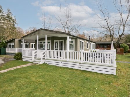 A single-story lodge with a white railing porch on a grassy yard at Cosy Corner in White Cross Bay near Troutbeck Bridge