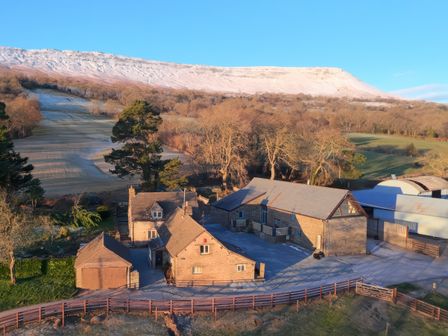A cluster of stone buildings with a wooded area and snow-covered hill in the background at White Haywood Barn in Craswall