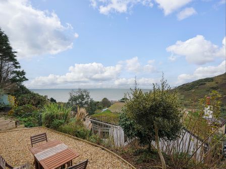 An outdoor seating area with a wooden table and chairs overlooking plants rooftops and the sea at Waves Edge in Looe