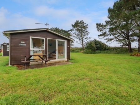 A small brown cabin with a picnic table outside on grass near trees at High Cove near St Merryn