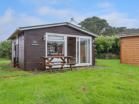 A small wooden cabin with sliding glass doors and a picnic table on a grassy lawn at Newtrain Bay near St Merryn