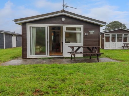 A wooden cabin with sliding glass door and window with curtains and a picnic table on the grass in front at Porthcothan Bay in St Merryn