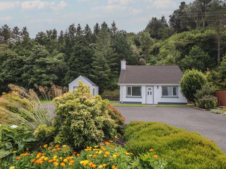 A white house with a brown roof surrounded by bushes and flowers in a forested area at Nelly's Chalet in Gortahork County Donegal
