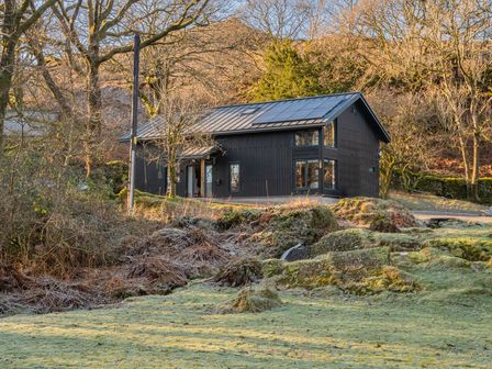A black wooden house with solar panels on the roof surrounded by grass and trees at Kolbeh Water Yeat near Coniston