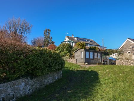 A garden with a wooden shed and a house on a hill surrounded by bushes at Swn Y Wylan in Red Wharf Bay near Benllech
