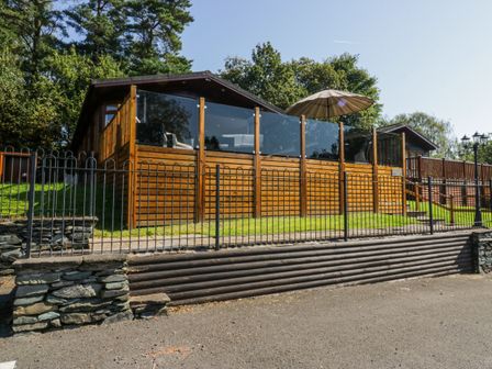 A wooden cabin with glass balcony panels and an outdoor umbrella behind a metal fence at Lakeland View Ambleside 86 White Cross Bay near Windermere