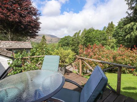 A wooden deck with a glass table and two cushioned chairs overlooking trees and hills at The Farmhouse in Coniston