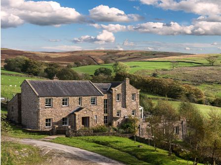 A stone house surrounded by green fields and trees under a blue sky at High Fellside Hall in Sedbergh
