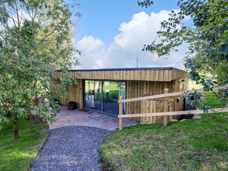 An outdoor view of a wooden lodge with sliding glass doors surrounded by grass trees and a gravel path at Orchard Lodge in Maerdy near Corwen