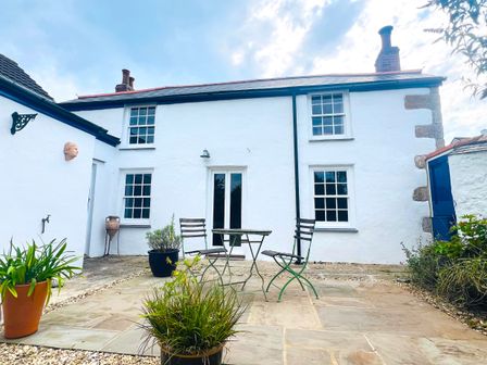 A patio with two chairs and a table in front of a white house with four windows and two chimneys at Rose Cottage in Falmouth