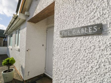 A front entrance with a white door and a sign reading The Gables on a textured wall with a small potted plant outside at The Gables in Tywyn